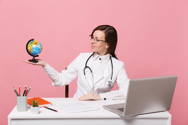 Médica sentar na mesa de trabalho no computador com documento médico segurar globo no hospital isolado no fundo da parede rosa pastel. Mulher de estetoscópio de óculos de vestido médico. Conceito de medicina de saúde.