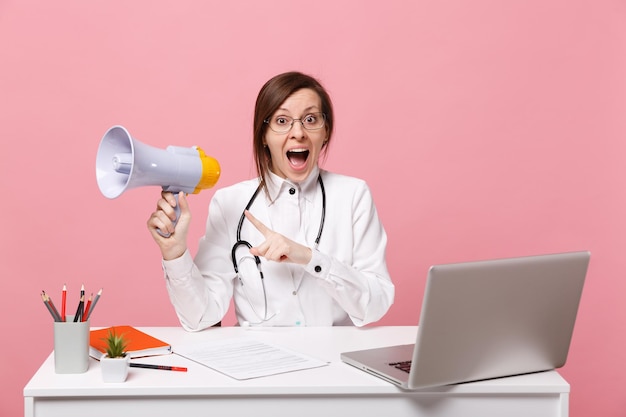 Médica sentar na mesa de trabalho no computador com documento médico segurar megafone no hospital isolado em fundo rosa pastel. Mulher de estetoscópio de óculos de vestido médico. Conceito de medicina de saúde.