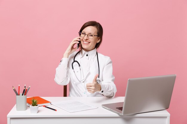 Médica sentar na mesa de trabalho no computador com documento médico segurar o celular no hospital isolado em fundo rosa pastel. mulher de estetoscópio de óculos de vestido médico. conceito de medicina de saúde.