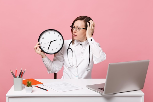 Médica sentar na mesa de trabalho no computador com documento médico segurar relógio no hospital isolado no fundo da parede rosa pastel. mulher de estetoscópio de óculos de vestido médico. conceito de medicina de saúde.