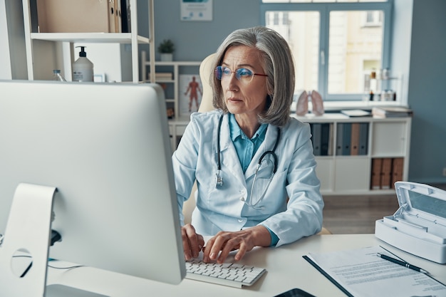 Médica séria e madura de jaleco branco, trabalhando usando o computador enquanto está sentada no escritório