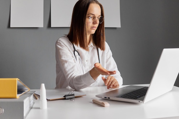 Médica séria trabalhando no escritório em seu local de trabalho olhando para o monitor do laptop falando com seu paciente pela internet tendo consulta médica on-line