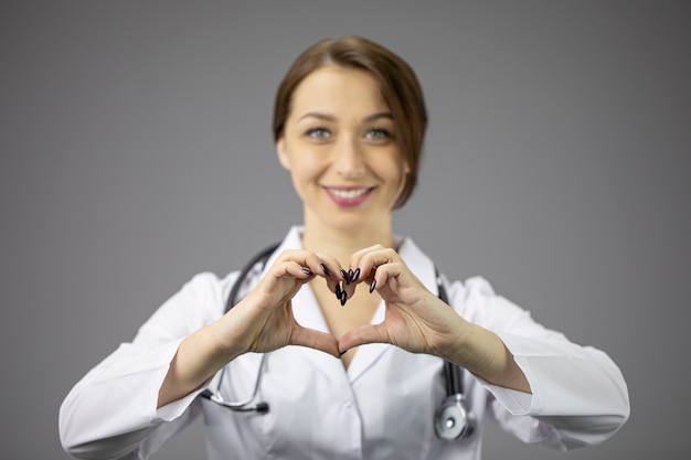 Médica sexy de jaleco branco na parede cinza mostra coração e sorrisos