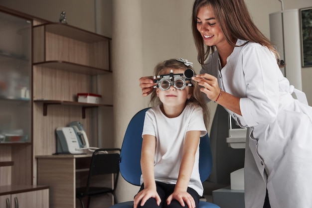 Foto médica sorridente atraente amando o trabalho dela. menina de óculos sentada na clínica e tendo seus olhos testados.