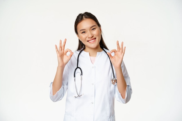 Médica sorridente, trabalhador de saúde asiático em uniforme de clínica médica, mostra sinais de ok, aprovar smth, dar permissão ao paciente, fica sobre fundo branco.