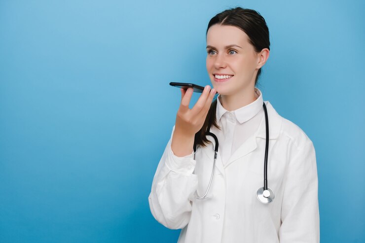 Médica sorridente, vestindo uniforme médico branco e estetoscópio segurando a fala do telefone, ativar o assistente de voz digital virtual no smartphone, consultar o paciente on-line, posando sobre a parede de fundo azul