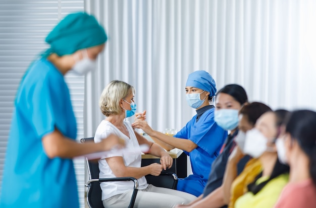 Médica usa máscara facial e uniforme do hospital, injetando vacina contra o coronavírus no ombro de uma mulher idosa, enquanto outras pacientes multinacionais esperam na fila em primeiro plano desfocado