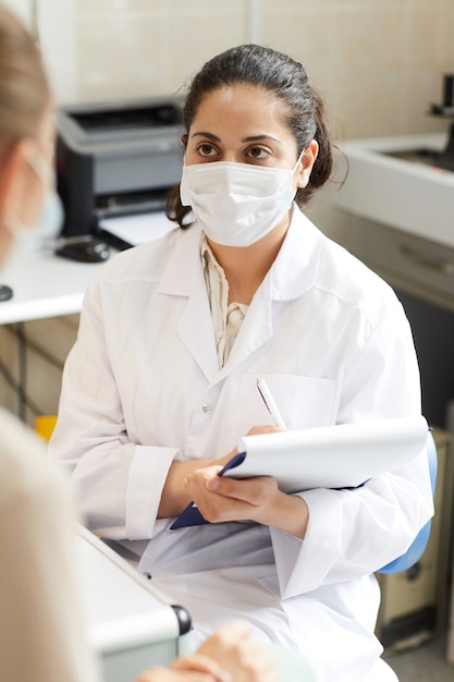 Médica usando máscara e jaleco branco prescrevendo receita para paciente no hospital