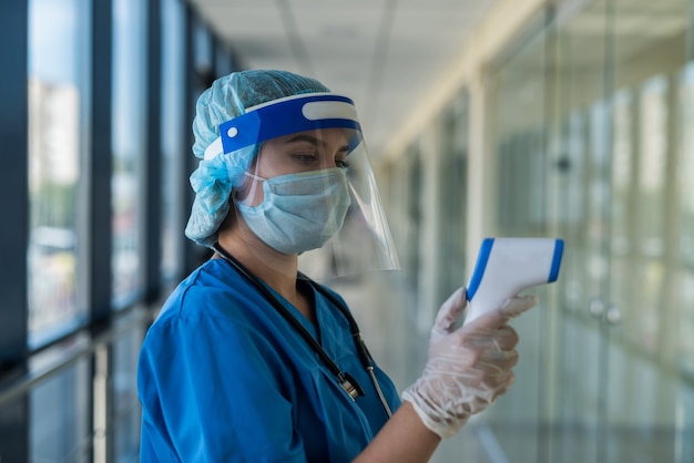 Médica usando máscara protetora e protetor facial, usando termômetro infravermelho para verificar a temperatura corporal na clínica. covid19