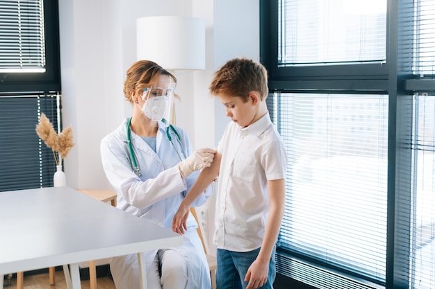 Médica vestindo uniforme branco e máscara de segurança aplicando gesso no ombro do menino após a injeção de vacinação pela janela