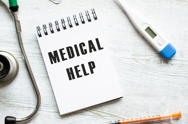MEDICAL HELP steht in einem Notizbuch auf einem hellen Holztisch neben einem Stethoskop.