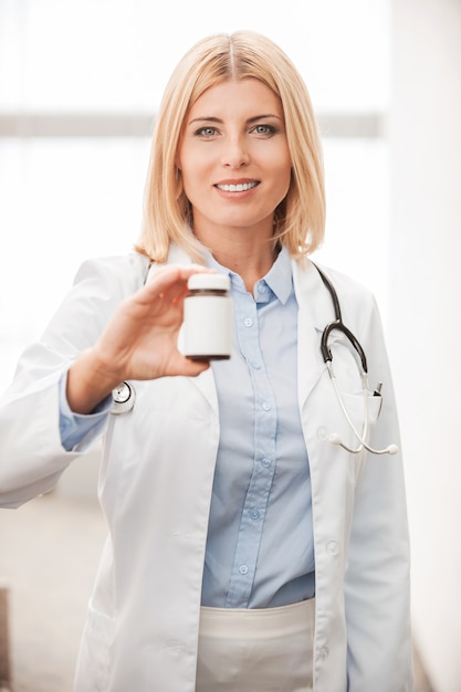 Medicamentos curativos. Confianza doctora en uniforme blanco sosteniendo un recipiente con medicamentos y sonriendo