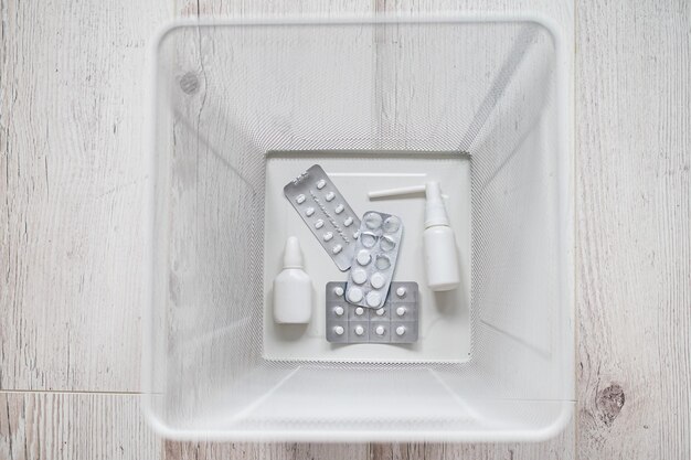 Medicamentos en una papelera de metal de malla blanca Vista superior Píldoras tiradas a la basura Una cesta de papel con medicamentos en el interior sobre un suelo de madera