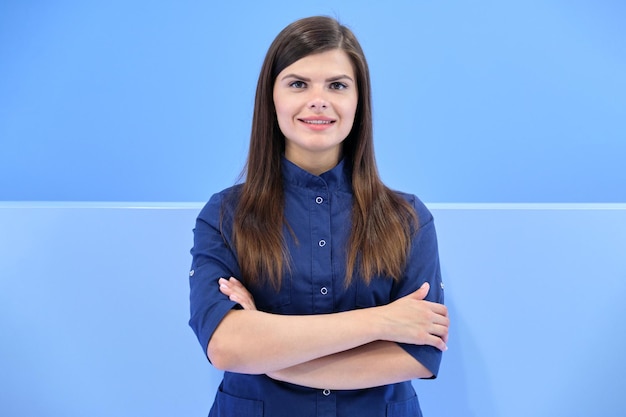 Medicina, beleza, odontologia, cosmetologia, cirurgia plástica, retrato de negócios confiante de jovem sorridente de uniforme azul com os braços cruzados, olhando para a câmera no fundo da clínica