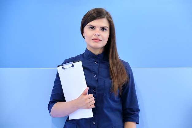 Medicina, belleza, odontología, cosmetología, cirugía plástica, retrato de negocios de mujer joven sonriente en uniforme azul con portapapeles mirando a cámara en el fondo de la clínica