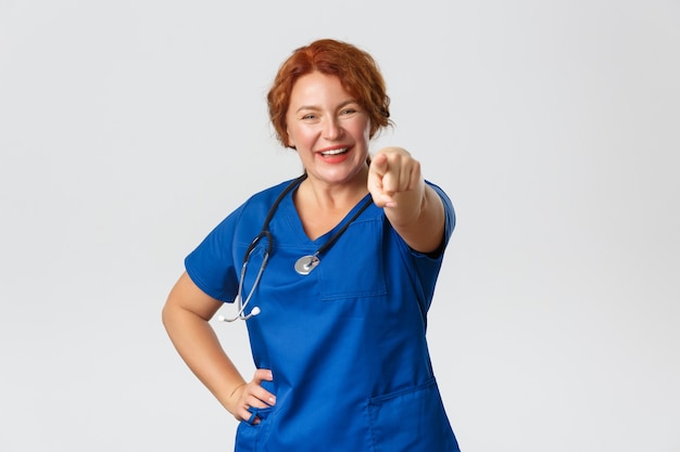 Medicina, concepto sanitario. Alegre sonriente y entusiasta médico de mediana edad, enfermera pelirroja apuntando con el dedo a la cámara
