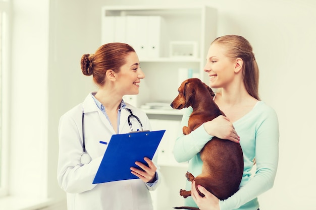 medicina, cuidados com animais de estimação e conceito de pessoas - mulher feliz segurando cachorro bassê e médico veterinário com prancheta na clínica veterinária