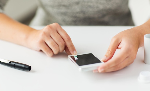 Medicina, diabetes, glucemia, atención de la salud y concepto de personas: cierre de las manos de la mujer con un teléfono inteligente que verifica el nivel de azúcar en la sangre con un glucómetro en casa