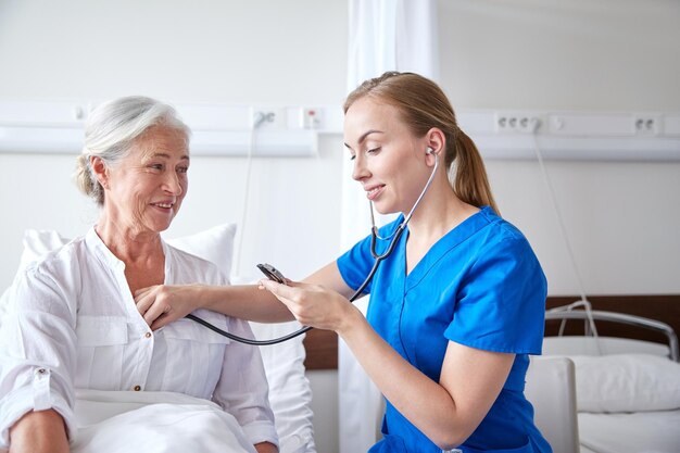 medicina, edad, apoyo, atención médica y concepto de personas - médico o enfermera con estetoscopio visitando a una anciana y revisando su latido cardíaco en la sala del hospital