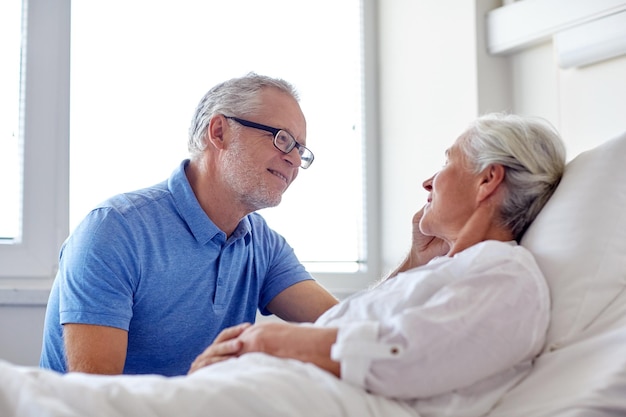 medicina, edad, apoyo, atención de la salud y concepto de la gente - anciano feliz visitando y animando a su mujer acostada en la sala del hospital