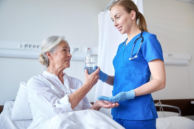 medicina, edad, atención de la salud y concepto de personas - enfermera que da medicamentos y un vaso de agua a una anciana en la sala del hospital