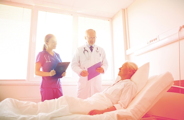 Medicina, edad, atención de la salud y concepto de personas - médico y enfermera con portapapeles visitando a una paciente mayor en la sala del hospital