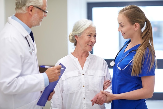 medicina, edad, atención de la salud y concepto de personas - médico masculino con portapapeles, enfermera joven y paciente anciana hablando en el pasillo del hospital