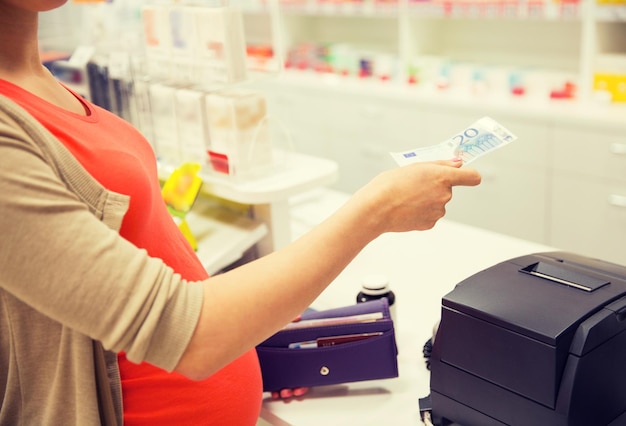 medicina, farmacêutica, cuidados de saúde e conceito de pessoas - close-up de mulher grávida dando dinheiro e comprando medicamentos na caixa registradora na farmácia