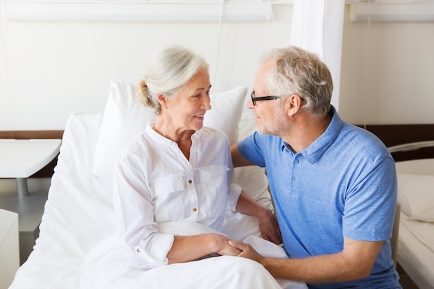 medicina, idade, apoio, cuidados de saúde e conceito de pessoas - feliz homem sênior visitando e torcendo sua mulher deitada na cama na enfermaria do hospital