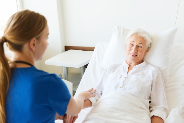 medicina, idade, apoio, cuidados de saúde e conceito de pessoas - médico ou enfermeiro visitando e torcendo mulher sênior deitada na cama na enfermaria do hospital