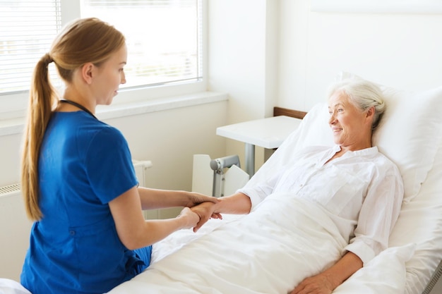 medicina, idade, apoio, cuidados de saúde e conceito de pessoas - médico ou enfermeiro visitando e torcendo mulher sênior deitada na cama na enfermaria do hospital