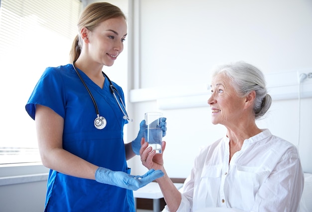 medicina, idade, cuidados de saúde e conceito de pessoas - enfermeira dando medicação e copo de água para mulher sênior na enfermaria do hospital