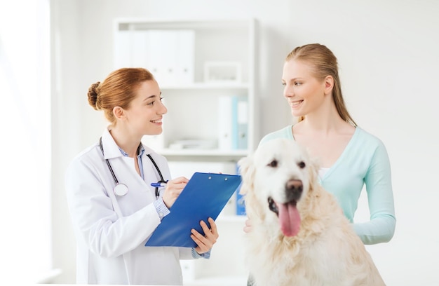 medicina, mascota, animales, atención de la salud y concepto de personas - mujer feliz con perro golden retriever y médico veterinario escribiendo en el portapapeles en la clínica veterinaria