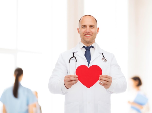 medicina, profissão, caridade e conceito de saúde - sorrindo médico masculino com coração vermelho e estetoscópio sobre grupo de médicos