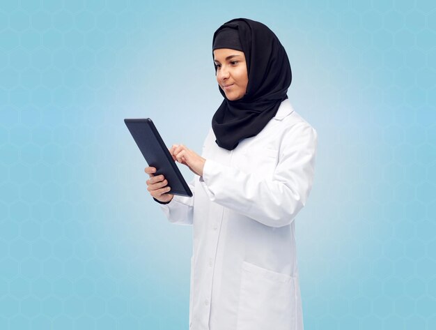 medicina, saúde, tecnologia e conceito de pessoas - sorridente médica muçulmana vestindo hijab e casaco branco com computador tablet sobre fundo azul