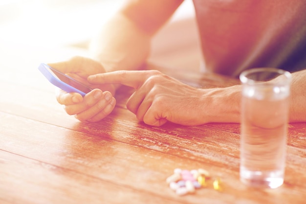 medicina, tecnología, suplementos nutricionales y conceptos de personas - primer plano de las manos masculinas teléfono inteligente, pastillas y agua en la mesa