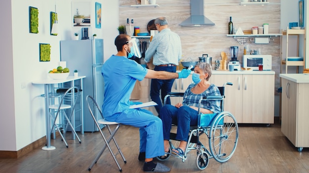 Medición de temperatura con termómetro infrarrojo en una mujer mayor discapacitada en silla de ruedas durante una visita domiciliaria en la pandemia de coronavirus. Trabajadora social con visera y máscara ayudando a la prevención del covid-19