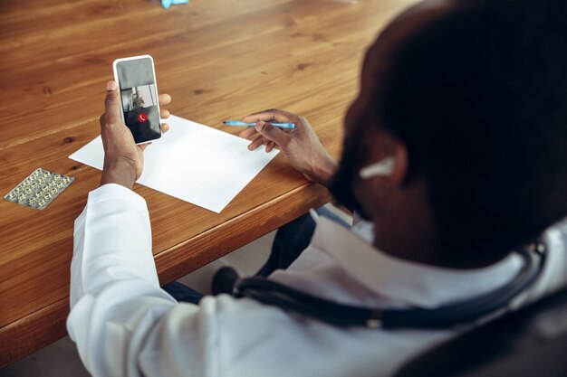 Médico aconselhando o paciente online com smartphone. médico afro-americano durante seu trabalho com pacientes, explicando receitas de remédios. trabalho árduo diário pela saúde e salvamento de vidas durante a epidemia.