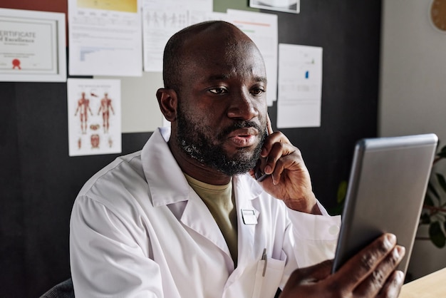 Médico africano con bata blanca sentado en su lugar de trabajo y sosteniendo una tableta frente a su cara