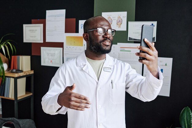 Médico africano con bata blanca usando teléfono móvil para consultas en línea durante el trabajo en la oficina