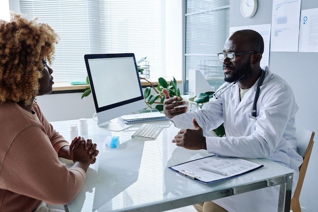 Médico africano de jaleco branco sentado à mesa e dando recomendações a mulheres africanas durante