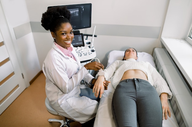 Médico africano jovem sorridente usando sonda de ultra-som no pulso do paciente jovem mulher caucasiana