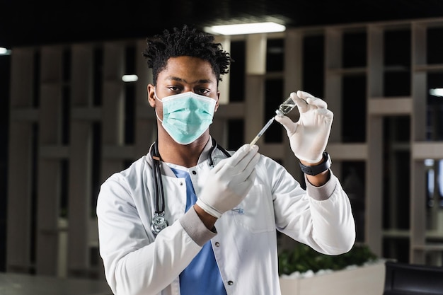 Médico africano con máscara médica se prepara para inyectar la vacuna coronavirus covid19 Médico negro con bata médica blanca con jeringa para vacunar