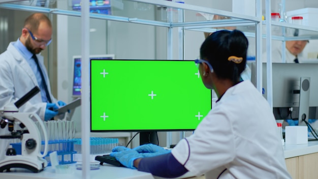 Médico africano que trabaja en la computadora con pantalla verde en un moderno laboratorio equipado. Equipo multiétnico de microbiólogos que realizan investigación de vacunas escribiendo en un dispositivo con clave de croma, pantalla de maqueta aislada.