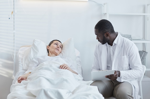 Médico africano con tarjeta médica hablando con el paciente que está acostado en la cama de un hospital