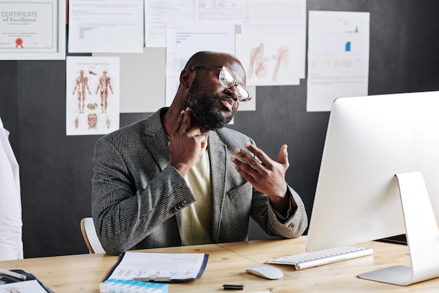 Médico africano tocando sua garganta e conversando com o paciente online no computador sentado no workpla