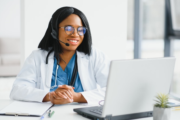 Médico africano usar fone de ouvido, consultar o paciente, fazer uma chamada de vídeo online com webcam na tela do laptop.