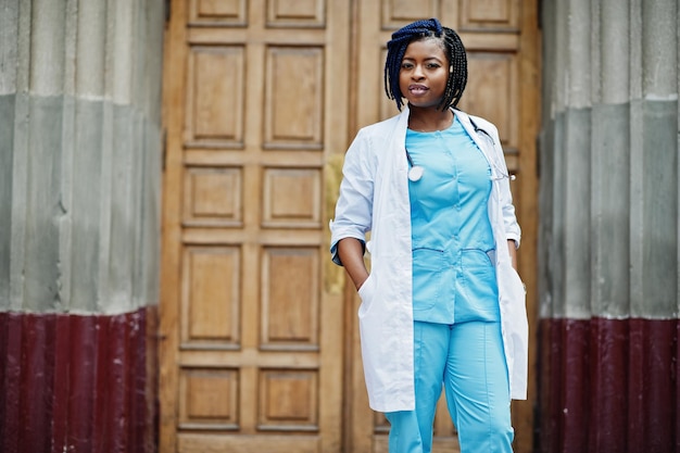 Médico afro-americano elegante com estetoscópio e jaleco posado contra a porta do hospital