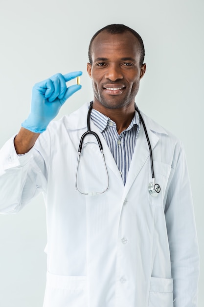 Médico afro-americano experiente e alegre segurando um comprimido enquanto se sente feliz