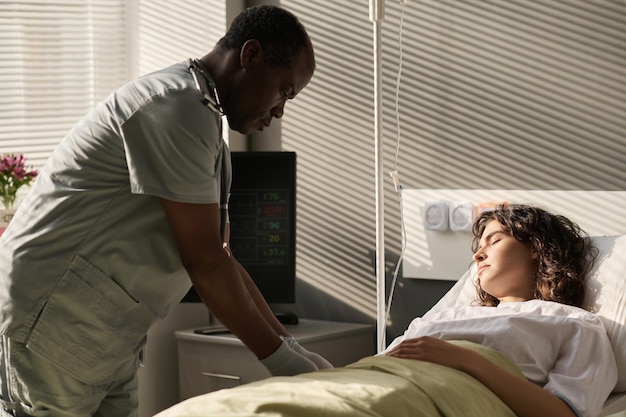 Médico afro-americano fazendo conta-gotas para jovem paciente enquanto ela dormia na cama na enfermaria